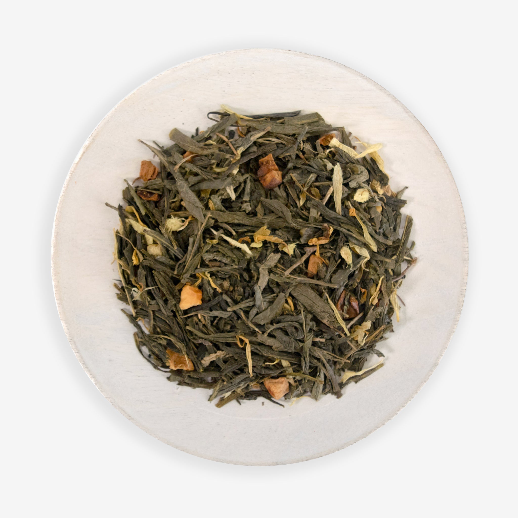 A top view of a bowl containing loose green tea leaves, with pieces of ginger and dried apple, against a white background.