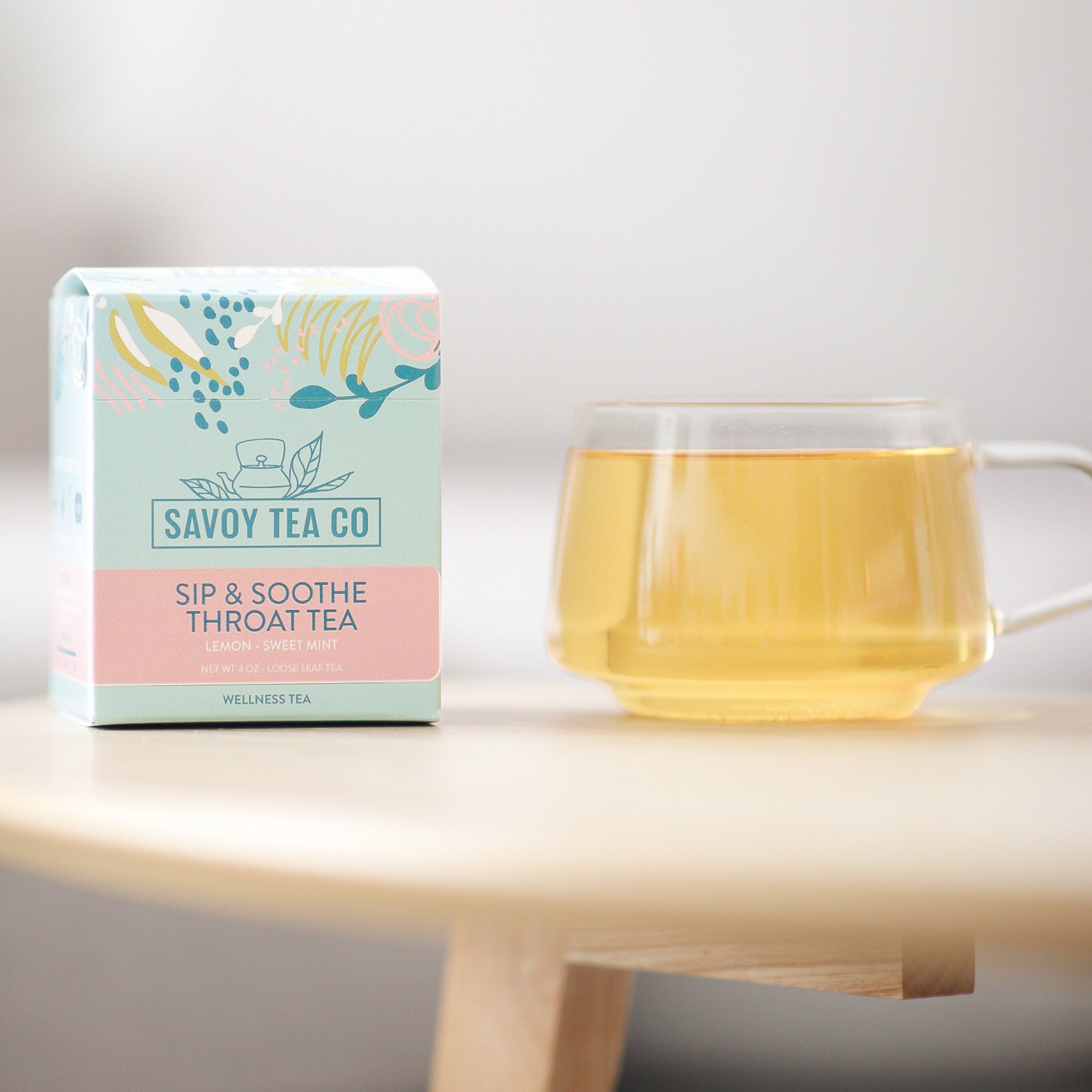A box of Savoy Tea Co. Sip & Soothe throat tea next to a clear mug filled with tea, placed on a wooden surface with a light background.